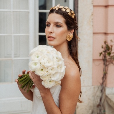 Mulher com vestido branco e acessórios dourados segurando ramo de flores brancas