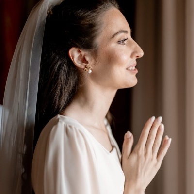 Noiva de perfil com vestido e véu branco, brincos dourados e pérola, mãos em oração