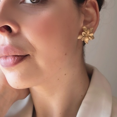 Brinco dourado em forma de flor usado por mulher com casaco bege claro e colar colorido