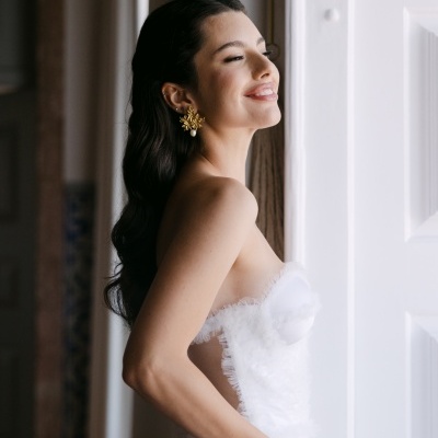 Mulher com vestido de noiva branco e brincos dourados ao lado de uma janela