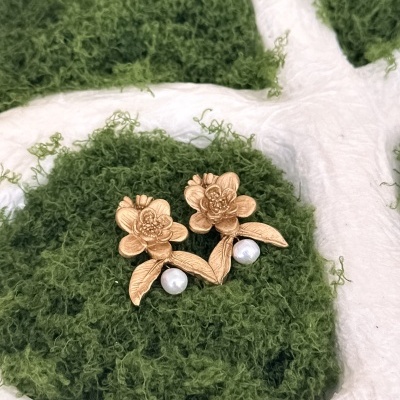 Brincos dourados em forma de flor com pérolas sobre fundo branco e musgo verde.