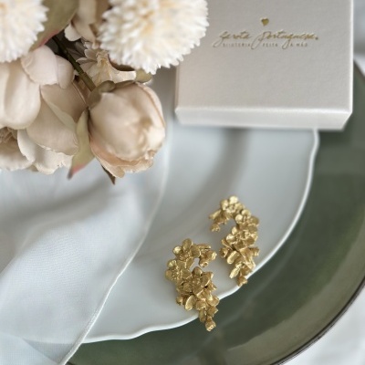 Brincos dourados florais sobre prato branco com embalagem ao fundo.
