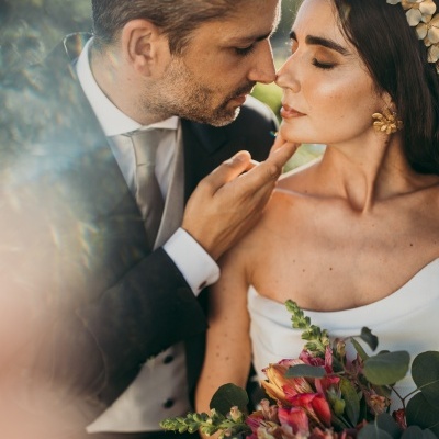 Casal de noivos em pose íntima, o homem com fato preto e a mulher com vestido branco e coroas de flores douradas segurando bouquet colorido