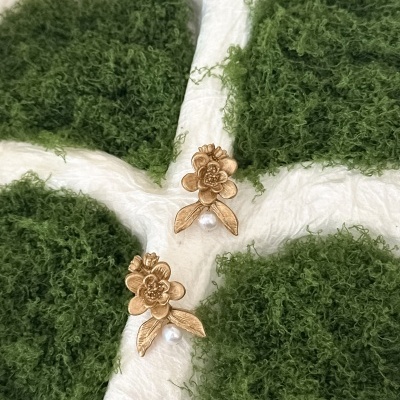 Brincos dourados em forma de flor com pérola sobre fundo branco com musgo verde.
