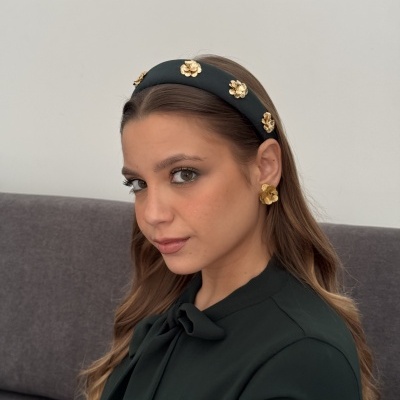 Mulher com faixa de cabelo preta com flores douradas e brincos dourados sentada num sofá cinzento