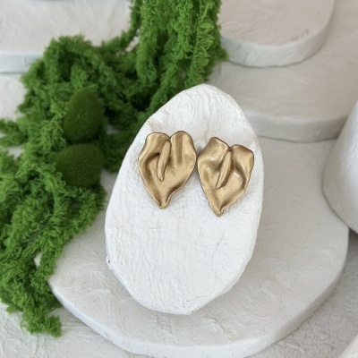 Brincos dourados em forma de folhas sobre suporte branco com plantas verdes ao lado