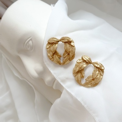 Brincos dourados em forma de folhas sobre tecido branco próximo a escultura de rosto humano