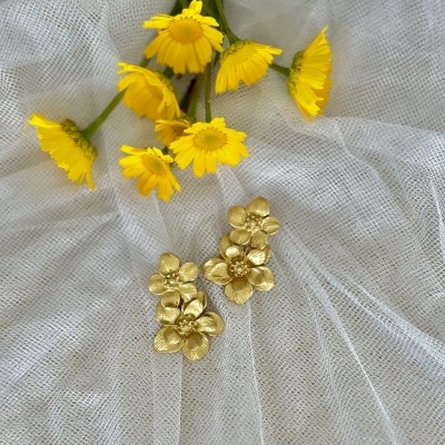 Brincos dourados de flores sobre tecido branco com flores amarelas ao fundo