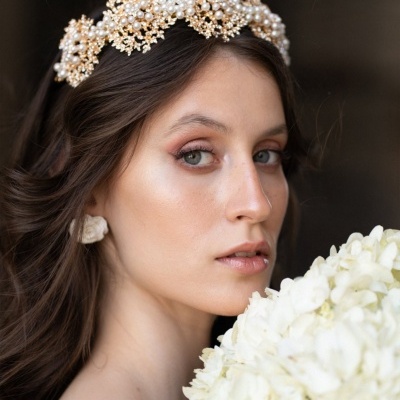 Mulher com tiara de pérolas e flores douradas, vestido branco e bouquet de flores brancas.