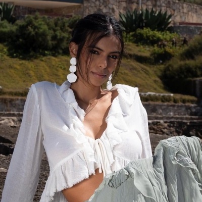 Mulher com blusa branca de tecido leve e brincos brancos largos, sentada em escadas de pedra