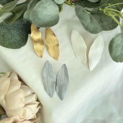 Três pares de brincos em forma de folha nas cores dourado, branco e prateado sobre tecido branco com folhas e flores decorativas