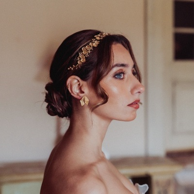 Mulher com fita dourada no cabelo e brincos dourados, top branco com padrão floral, parede e porta ao fundo