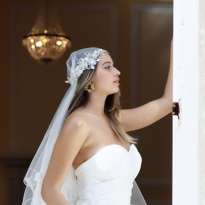 Mulher com vestido e véu de noiva branco, rosto de perfil e brincos dourados, candelabro desfocado ao fundo.