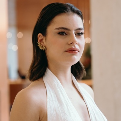 Mulher com vestido branco e brincos dourados, cabelo castanho ao ombro, em ambiente interior desfocado.