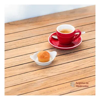 Copo de café expresso vermelho com pires e bolo pequeno numa tigela branca sobre mesa de madeira.