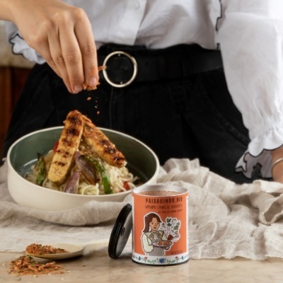 Lata de tempero PAISAGINDO.BIO à frente de tigela com noodles, legumes e tofu, mão a polvilhar tempero.