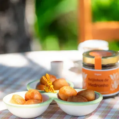 Taça branca com pãezinhos dourados e coelhinho amarelo em cerâmica no centro, mesa com toalha quadriculada e frasco ao fundo