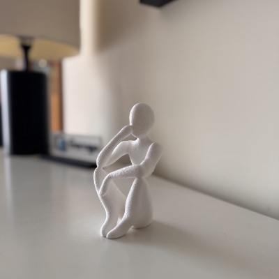 Estatueta branca abstrata de ser humano sentado sobre superfície branca com lâmpada preta ao fundo