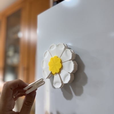 Imã de frigorífico flor branca com centro amarelo e mãos segurando objeto branco