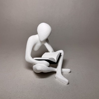 Estatueta branca de figura humana lendo um livro sobre fundo cinzento.