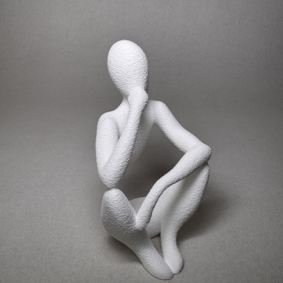 Estátua decorativa branca texturizada de figura humana sentada com a mão no rosto sobre fundo cinzento
