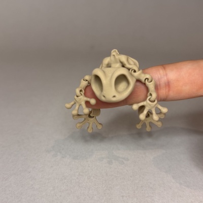 Figura pequena de rã flexível bege sobre dedo humano, fundo cinzento