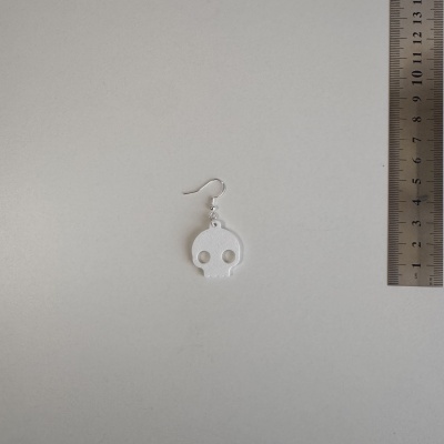 Brinco caveira branco com gancho prateado ao lado de régua metálica.