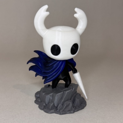 Boneco com cabeça branca, olhos negros e manto azul em base cinzenta tipo pedra