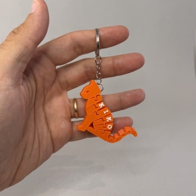 Chaveiro dinossauro laranja com letras brancas pendurado numa corrente metálica prateada segurado por mão com anel dourado