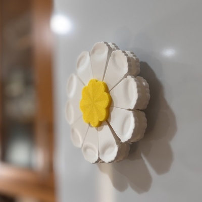 Magneto em forma de flor com pétalas brancas e centro amarelo numa superfície metálica cinza