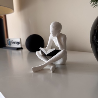 Figura decorativa branca a ler livro preto e branco sobre mesa branca com esfera preta e placa Garuga