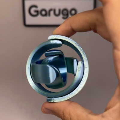 Peça metálica azul esverdeada com design circular e hélice, mão segurando, fundo cinzento com texto Garugo
