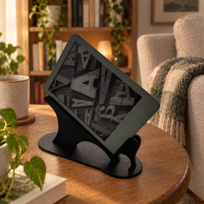 Tablet cinzento em suporte preto numa mesa de madeira com caneca, planta e livros, numa sala acolhedora com estante e sofá.