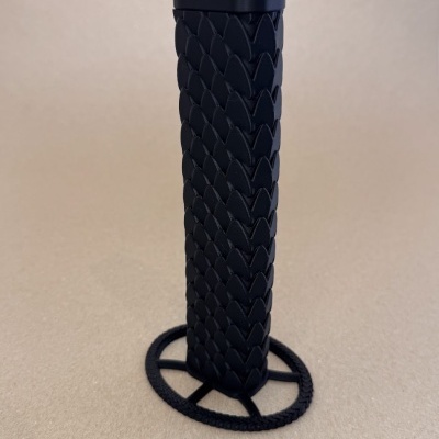 Objeto preto cilíndrico com padrão de escamas e base circular sobre superfície castanha clara