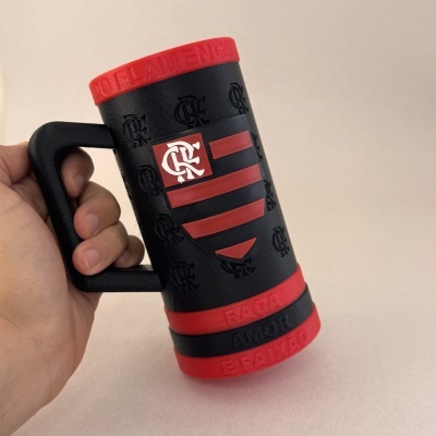 Caneca preta e vermelha com logotipo Flamengo e texto em relevo, segurada por mão.
