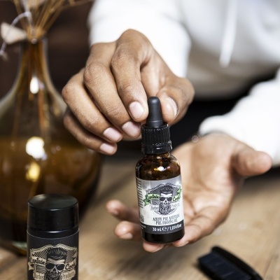 Produtos de barbear Captain Cook incluindo óleo pré-afeitar e pó em frascos sobre mesa de madeira com escova preta e vaso de flores secas ao fundo.