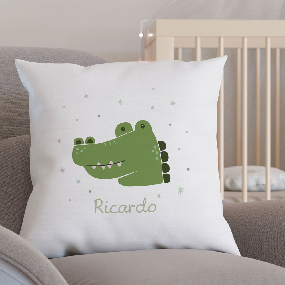 Almofada branca com crocodilo verde e nome Ricardo, sobre cadeira cinza, com berço ao fundo