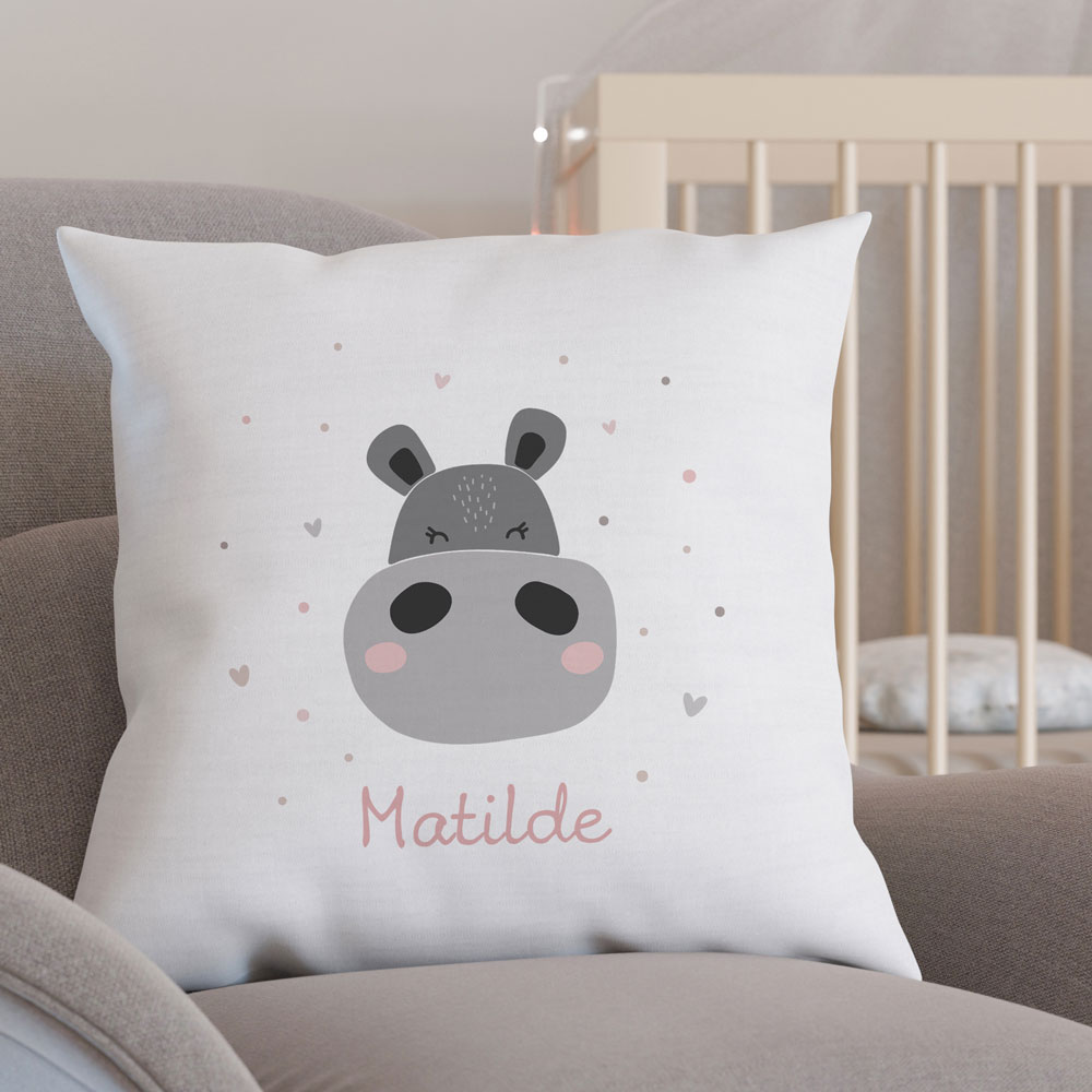 Almofada branca com estampa de hipopótamo e nome Matilde numa cadeira cinzenta perto de berço creme.