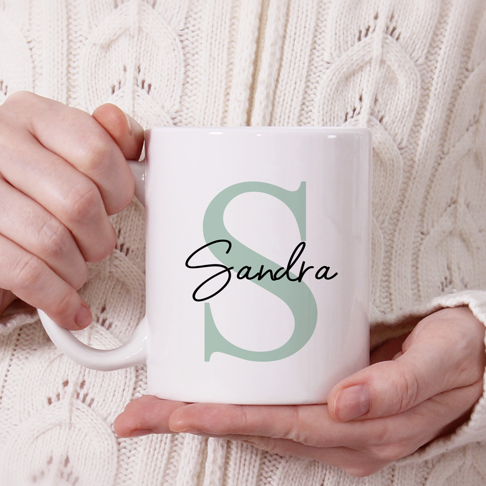 Caneca branca com a letra S verde e o nome Sandra preto, segurada com mãos sobre suéter branco em malha