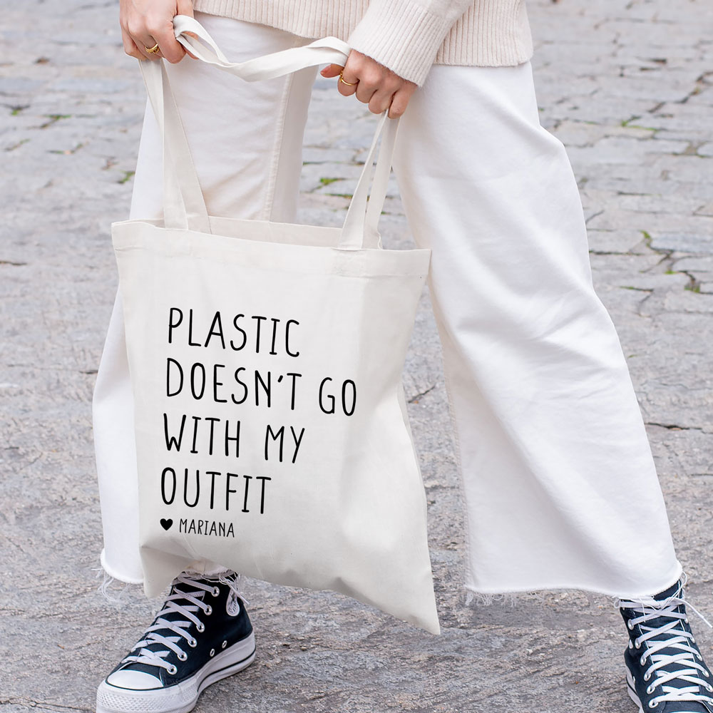 Saco de tecido branco com texto a preto segurado por pessoa com calças brancas e ténis pretos em chão de pedra