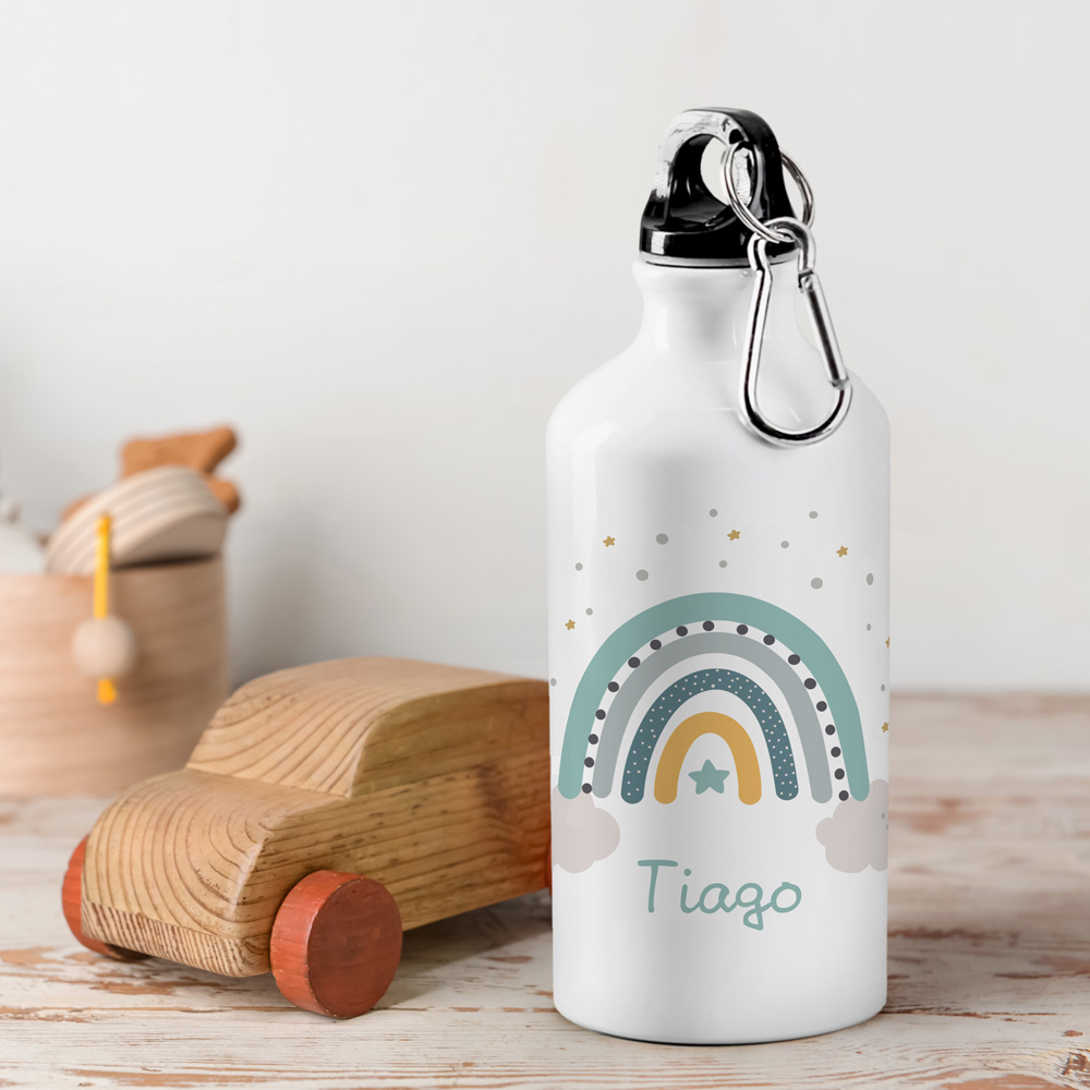 Garrafa de alumínio branca com desenho de arco-íris colorido e nome 'Tiago', junto a um brinquedo de carro de madeira.