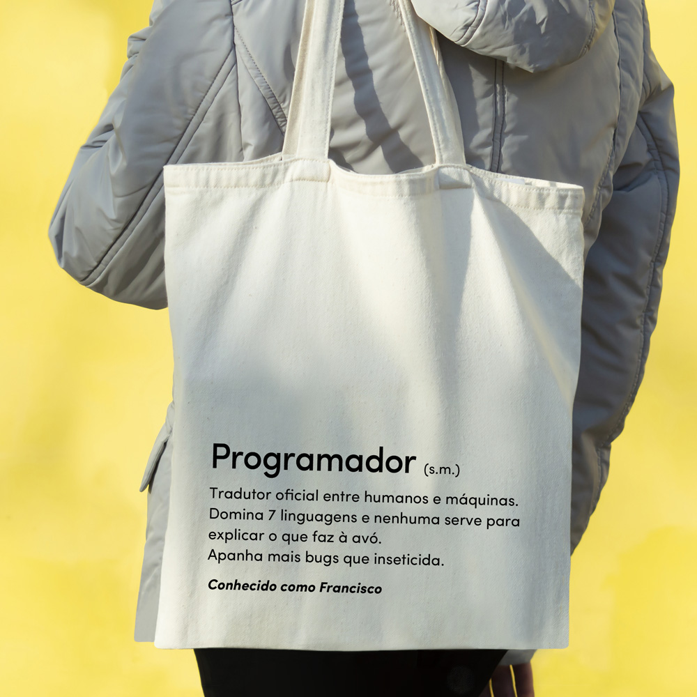 Sacola de tecido cru com texto em português, carregada ao ombro, contra fundo amarelo.