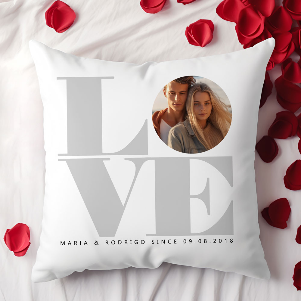 Almofada branca com palavra LOVE e foto de casal, sobre cama com pétalas de rosa vermelhas.