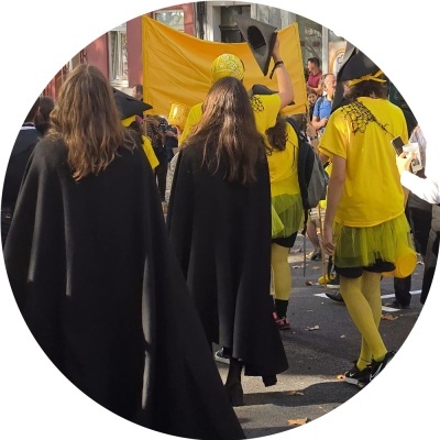 Pessoas em desfile trajando capas pretas e camisolas amarelas com acessórios de carnaval.