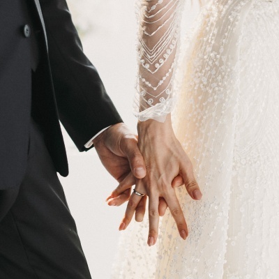 Mãos dadas de um casal, homem com fato preto e mulher com vestido de casamento branco bordado