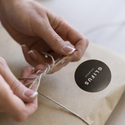 Pacote de papel castanho a ser amarrado com corda rústica por mãos femininas.