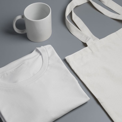 T-shirt branca, caneca branca e bolsa de tecido branca sobre fundo cinzento