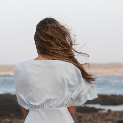 Pessoa de costas com vestido branco e cabelo ao vento junto ao mar