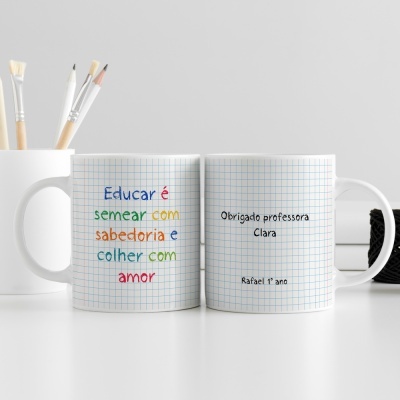 Duas canecas brancas quadriculadas com textos coloridos e pretos sobre agradecimento a professora, sobre mesa branca com pincéis e bola de linha.