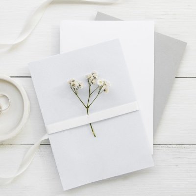 Convites brancos com flores secas, alianças num prato branco e fita branca sobre madeira